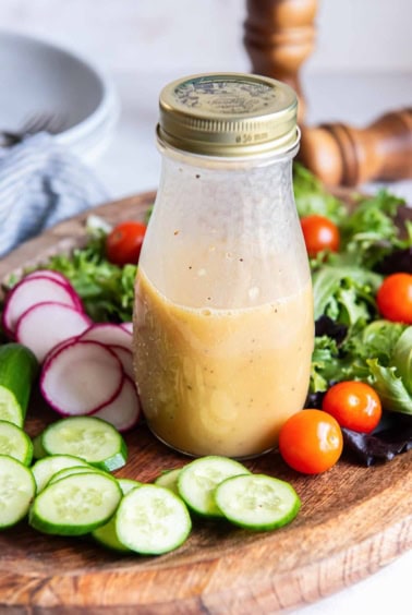 A jar of apple cider vinaigrette on a wood platter with salad ingredients.
