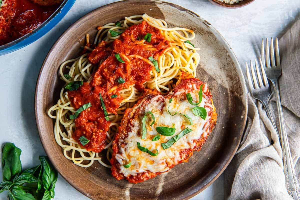 A plate of spaghetti topped with marinara sauce is served alongside a piece of chicken parmesan that is covered in melted cheese and garnished with fresh basil. A fork and knife are placed on a beige napkin at the side. Fresh basil leaves are in the corner.