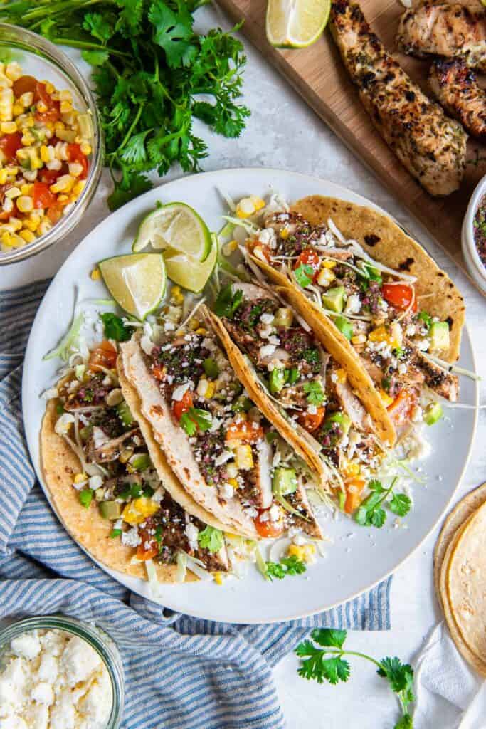Chimichurri chicken tacos with corn, tomatoes, avocado, and cabbage on a white plate.