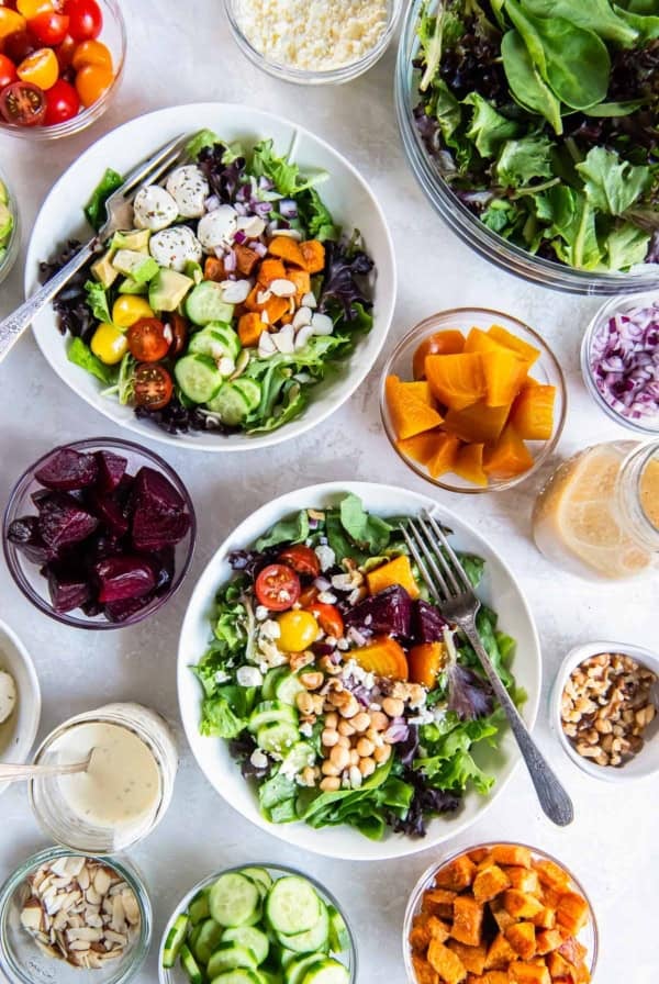 Two green salads in white bowls next to salad ingredients in small bowls.
