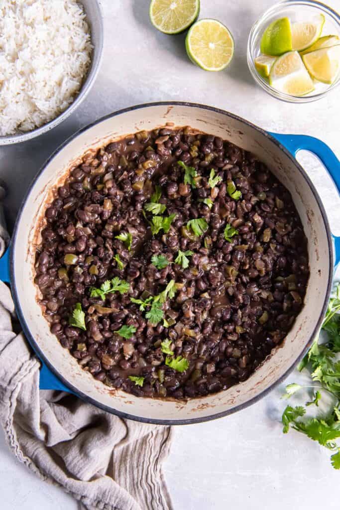 Cuban black beans in a skillet next to a bowl of rice and lime wedges.