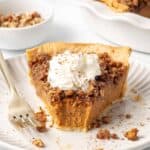 A slice of praline pumpkin pie topped with whipped cream with a bite missing.