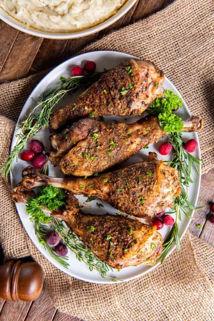 Roasted turkey drumsticks on a white platter with fresh herbs and cranberries.