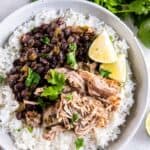 Cuban pork on white rice with black beans in a white bowl next to cilantro and limes.