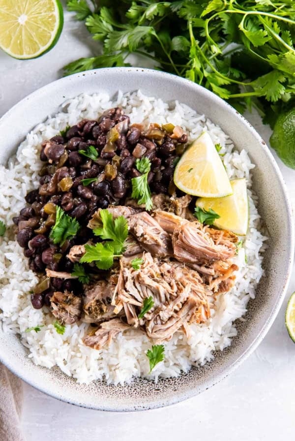 Cuban pork on white rice with black beans in a white bowl next to cilantro and limes.