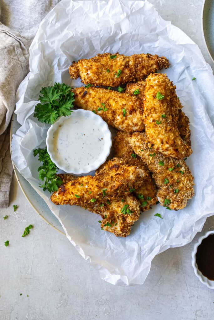 Air fryer chicken tenders on parchment paper with sprigs of parsley and Ranch in a small bowl.