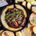 Crostini and apple slices surrounding baked brie with jam in a cast iron skillet.