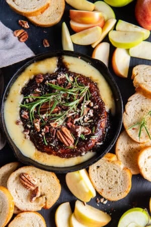 Crostini and apple slices surrounding baked brie with jam in a cast iron skillet.