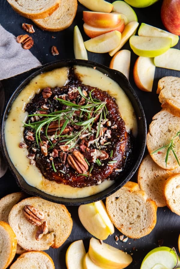 Crostini and apple slices surrounding baked brie with jam in a cast iron skillet.