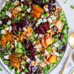 A roasted beet salad on a white platter with serving spoons lying next to it.