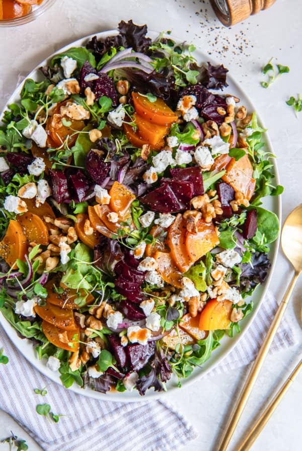 A roasted beet salad on a white platter with serving spoons lying next to it.