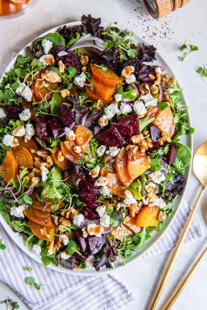 A roasted beet salad on a white platter with serving spoons lying next to it.