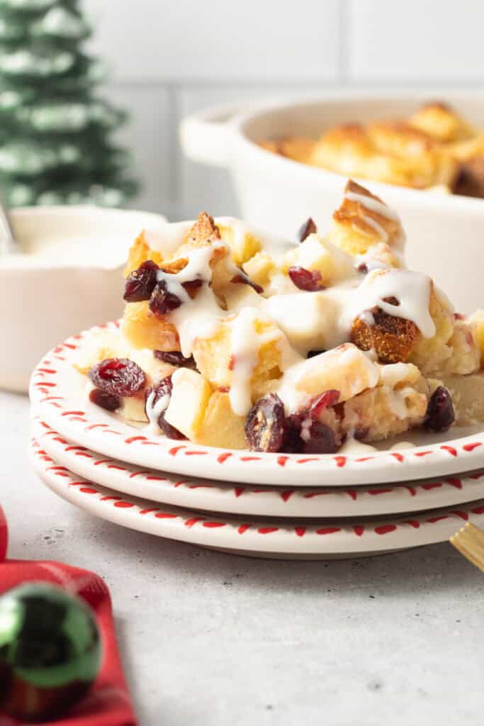 A plate with a serving of Christmas bread pudding with apples and cranberries topped with rum cream sauce on a kitchen counter.