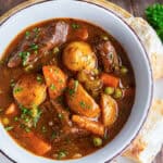 A bowl of beef stew resting on a white plate with a spoon and slices of bread.