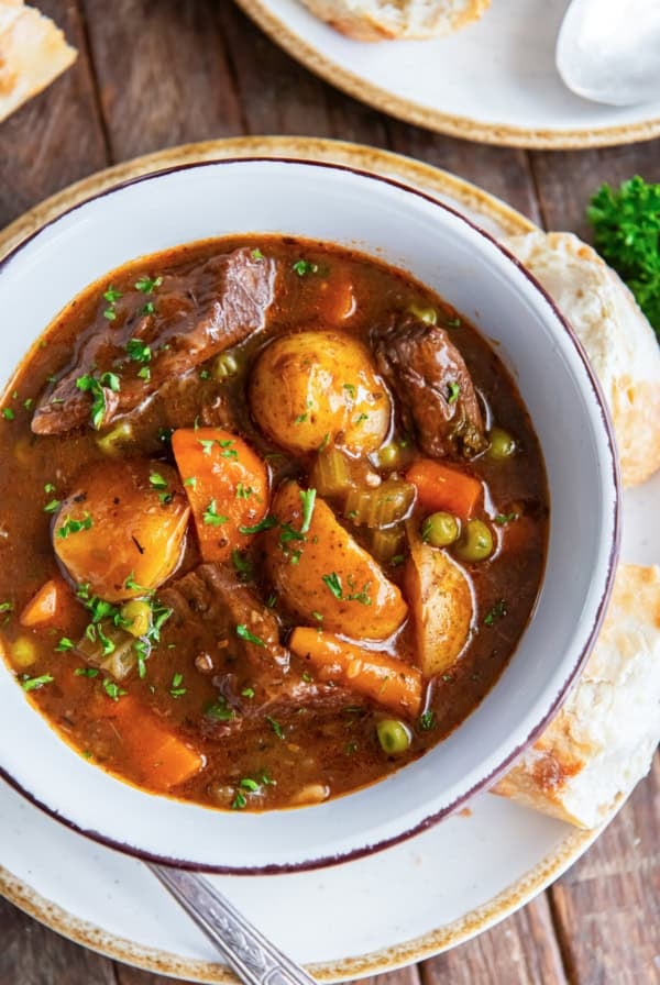 A bowl of beef stew resting on a white plate with a spoon and slices of bread.