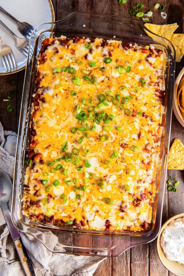 Chili cornbread casserole topped with green onions on a brown board next to a bowl of sour cream and tortilla chips.