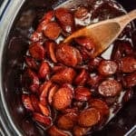 A wooden spoon stirring BBQ cocktail sausages in a crock pot.