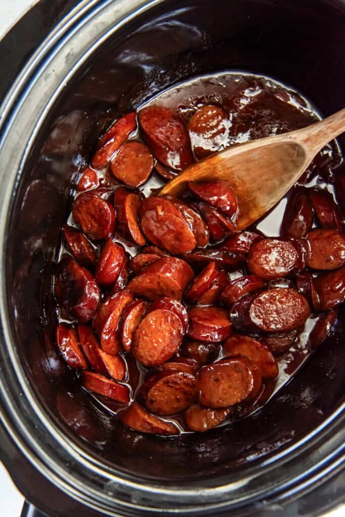 A wooden spoon stirring BBQ cocktail sausages in a crock pot.