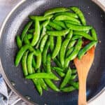 Sugar snap peas in a skillet with a wooden spatula.