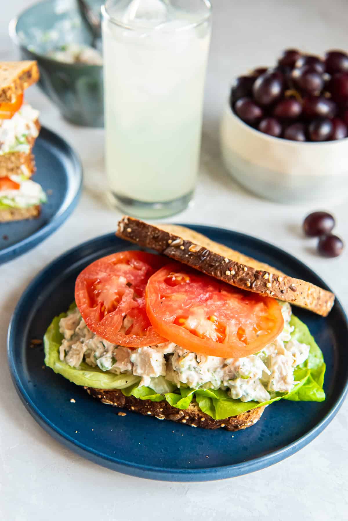 Chicken salad on toasted wheat bread with lettuce and tomato on a blue plate in front of a glass of lemonade.