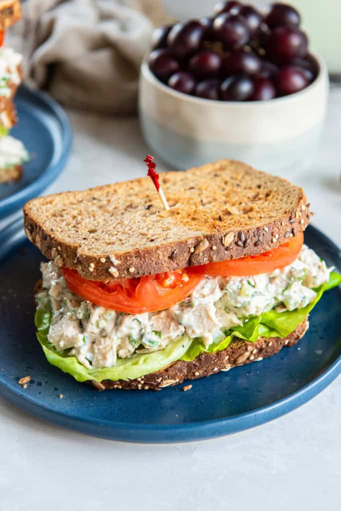 A chicken salad sandwich with lettuce and tomato on a blue plate in front of a bowl of grapes.