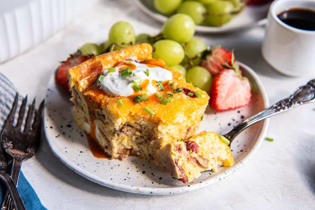 A slice of quiche casserole topped with sour cream and hot sauce on a white plate with a fork.