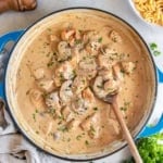 A wooden spoon resting in a pan of chicken stroganoff next to a bowl of egg noodles.