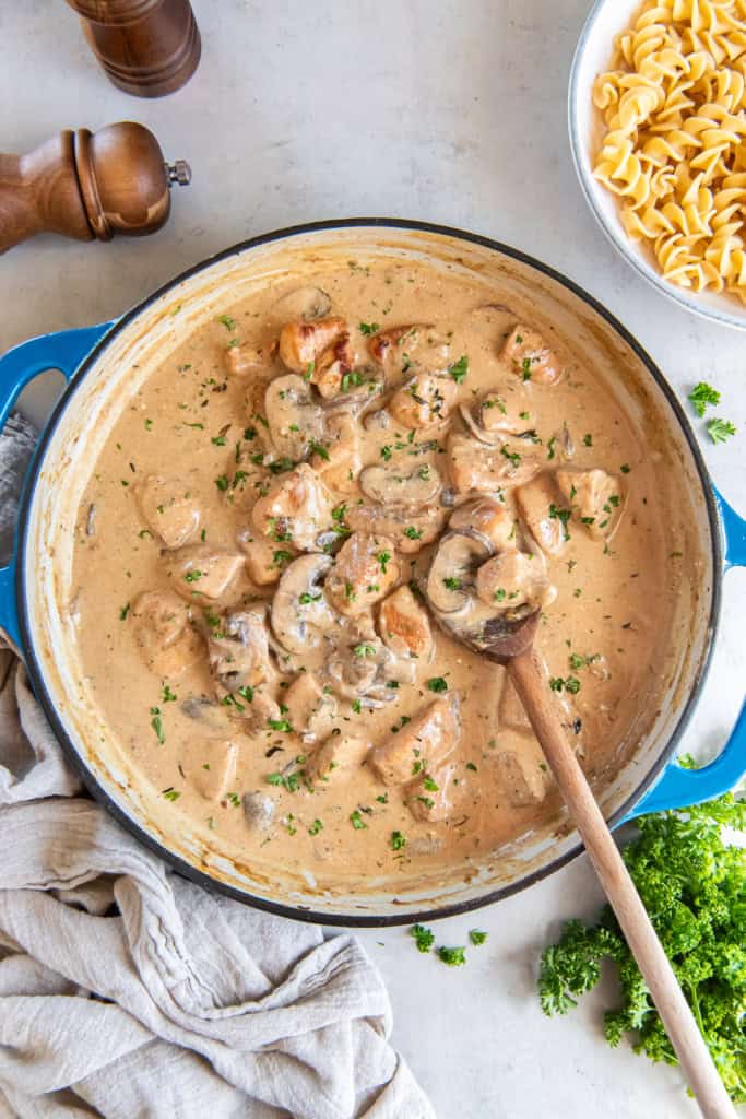 A wooden spoon resting in a pan of chicken stroganoff next to a bowl of egg noodles.
