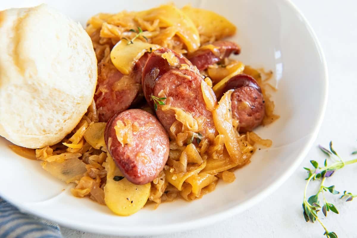 A serving of kielbasa and sauerkraut in a white bowl with a dinner roll.