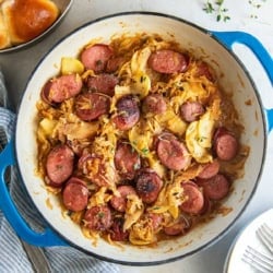 Kielbasa and sauerkraut with apples in a skillet next to a pan of dinner rolls.