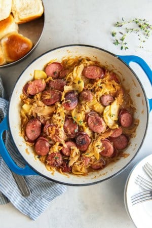 Kielbasa and sauerkraut with apples in a skillet next to a pan of dinner rolls.