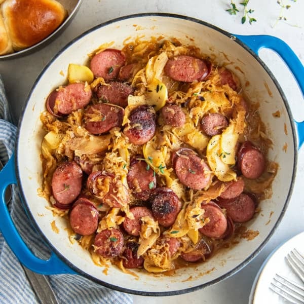 Kielbasa and sauerkraut with apples in a skillet next to a pan of dinner rolls.