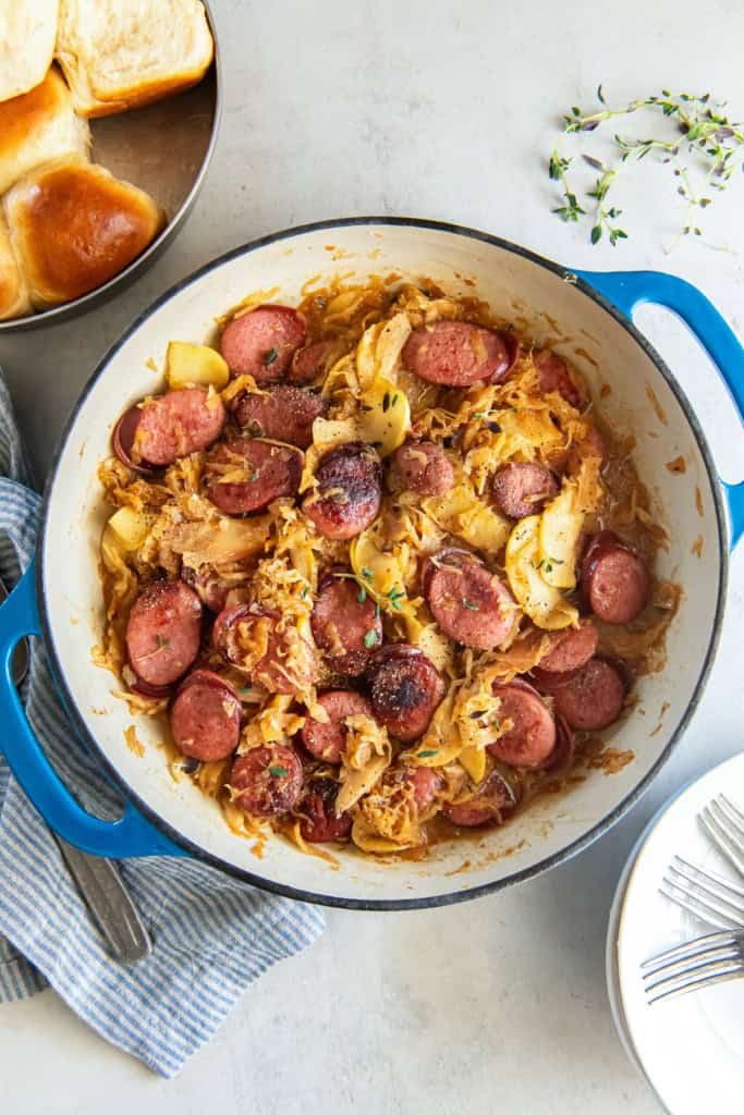 Kielbasa and sauerkraut with apples in a skillet next to a pan of dinner rolls.