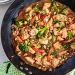 A chicken and vegetable stir fry with peanut sauce in a cast iron skillet next to a bowl of rice and green kitchen towel.