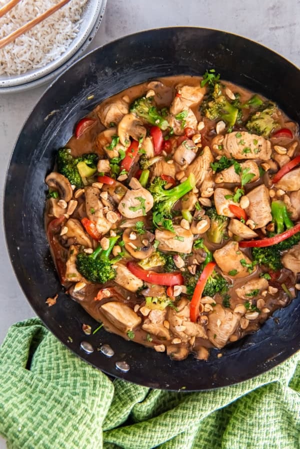 A chicken and vegetable stir fry with peanut sauce in a cast iron skillet next to a bowl of rice and green kitchen towel.