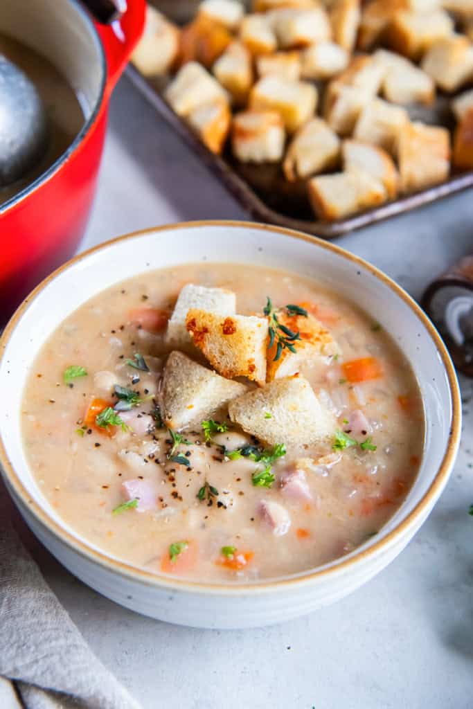 A bowl of white bean and ham soup topped with croutons next to a pot of soup and more croutons on a baking sheet.