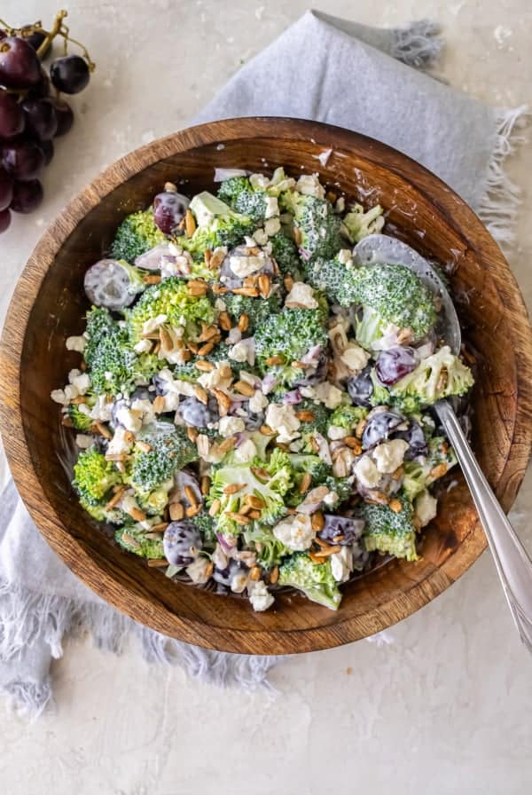 A serving spoon resting in a wood bowl filled with broccoli grape salad.