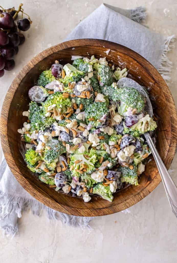 A serving spoon resting in a wood bowl filled with broccoli grape salad.