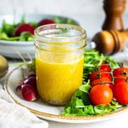 A mason jar filled with champagne vinaigrette on a plate with salad greens and cherry tomatoes.