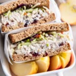 A lightened up chicken saad sandwich on wheat bread sliced in half in small ceramic dish with sliced apple.
