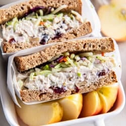 A lightened up chicken saad sandwich on wheat bread sliced in half in small ceramic dish with sliced apple.