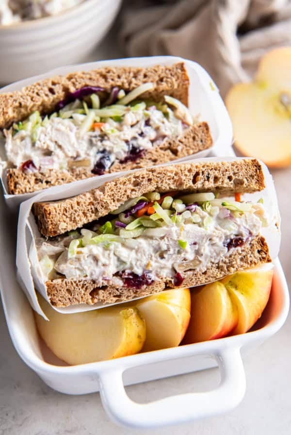 A lightened up chicken saad sandwich on wheat bread sliced in half in small ceramic dish with sliced apple.