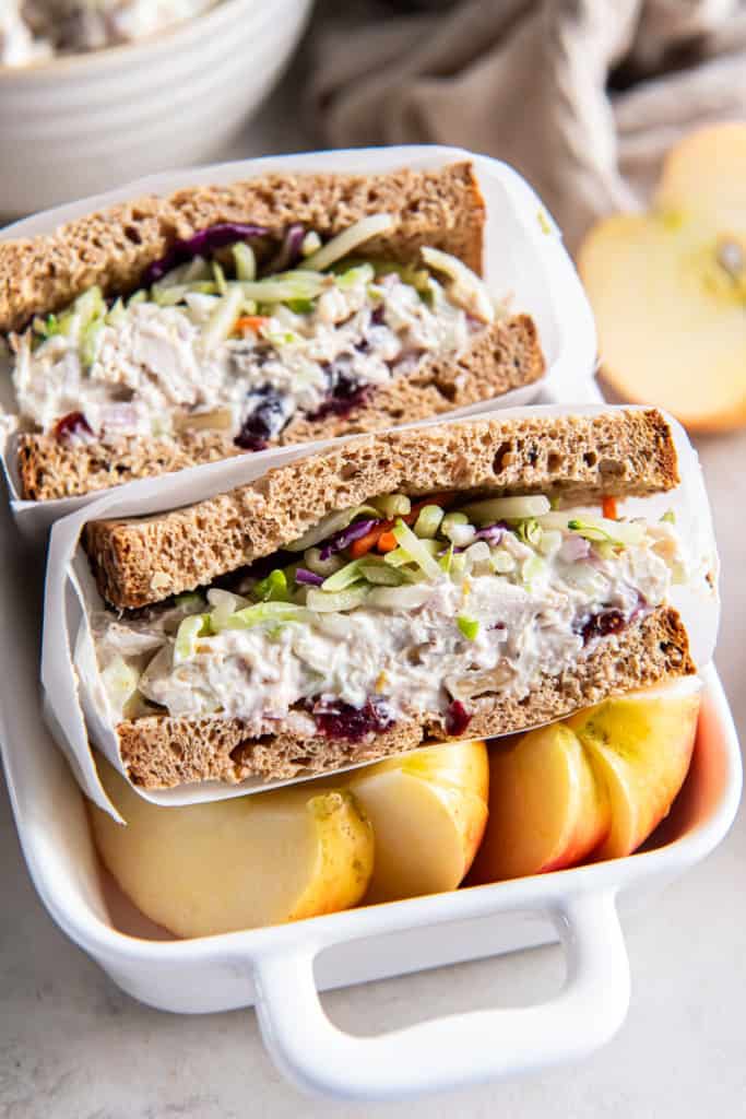 A lightened up chicken saad sandwich on wheat bread sliced in half in small ceramic dish with sliced apple.