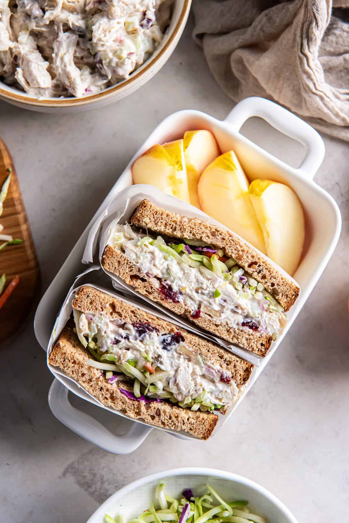 A halved chicken salad sandwich in parchment paper in a small ceramic dish with sliced apples.
