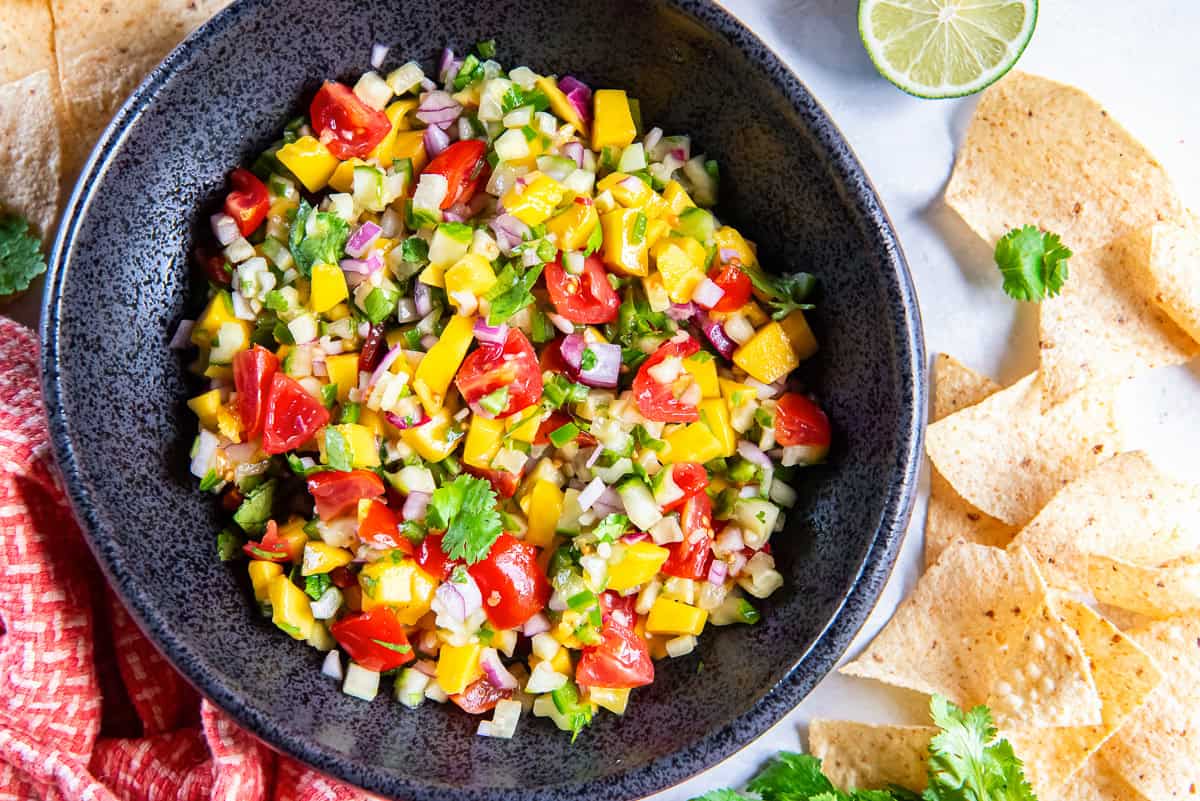 A bowl of mango cucumber salsa next to an orange and white kitchen cloth and tortilla chips.