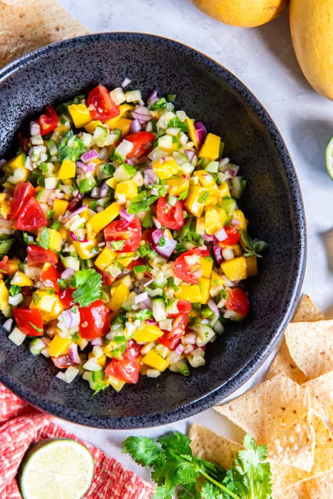Mango cucumber salsa in a black serving bowl with tortilla chips scattered around it.