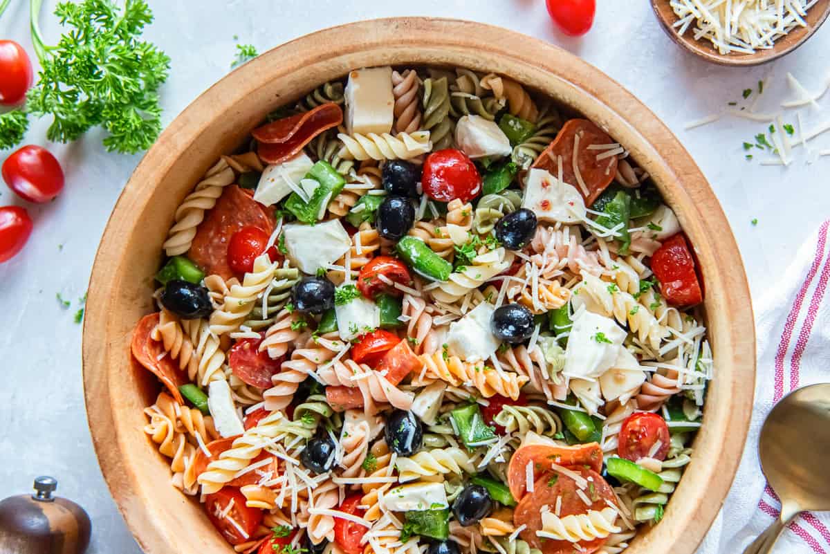 Pizza Pasta Salad 5 A wood bowl filled with pizza pasta salad surrounded by cherry tomatoes and fresh herbs.