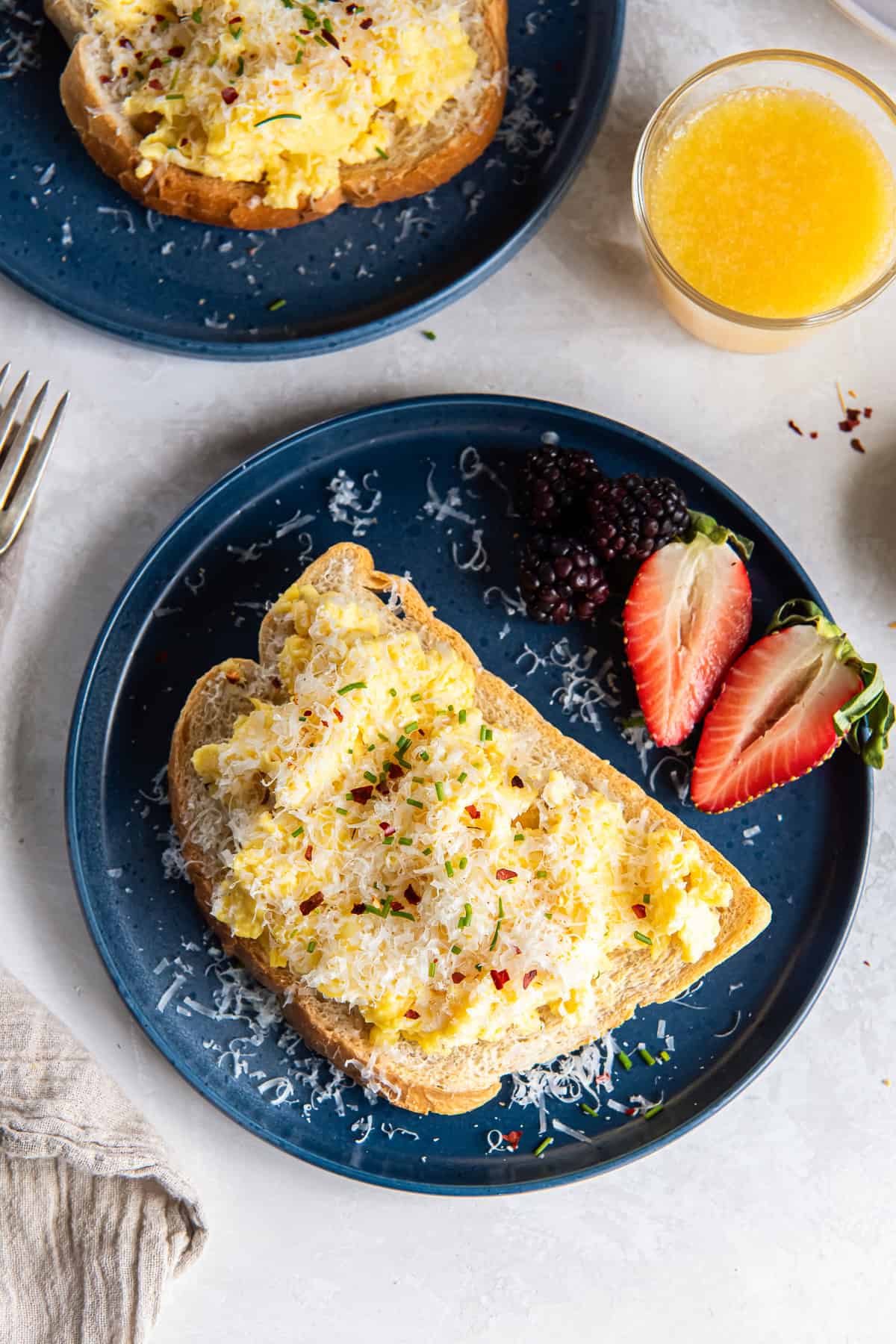 Two blue plates with toast topped with ricotta scrambled eggs next to a glass of orange juice.