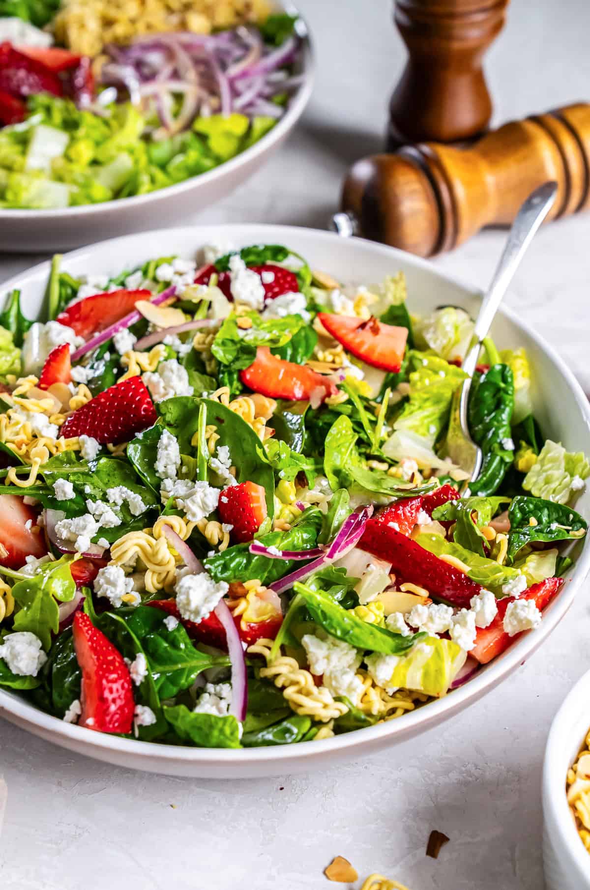 A fork resting in a bowl of strawberry crunch salad with greens, ramen noodles and champagne vinaigrette with salt and pepper shakers in the background.