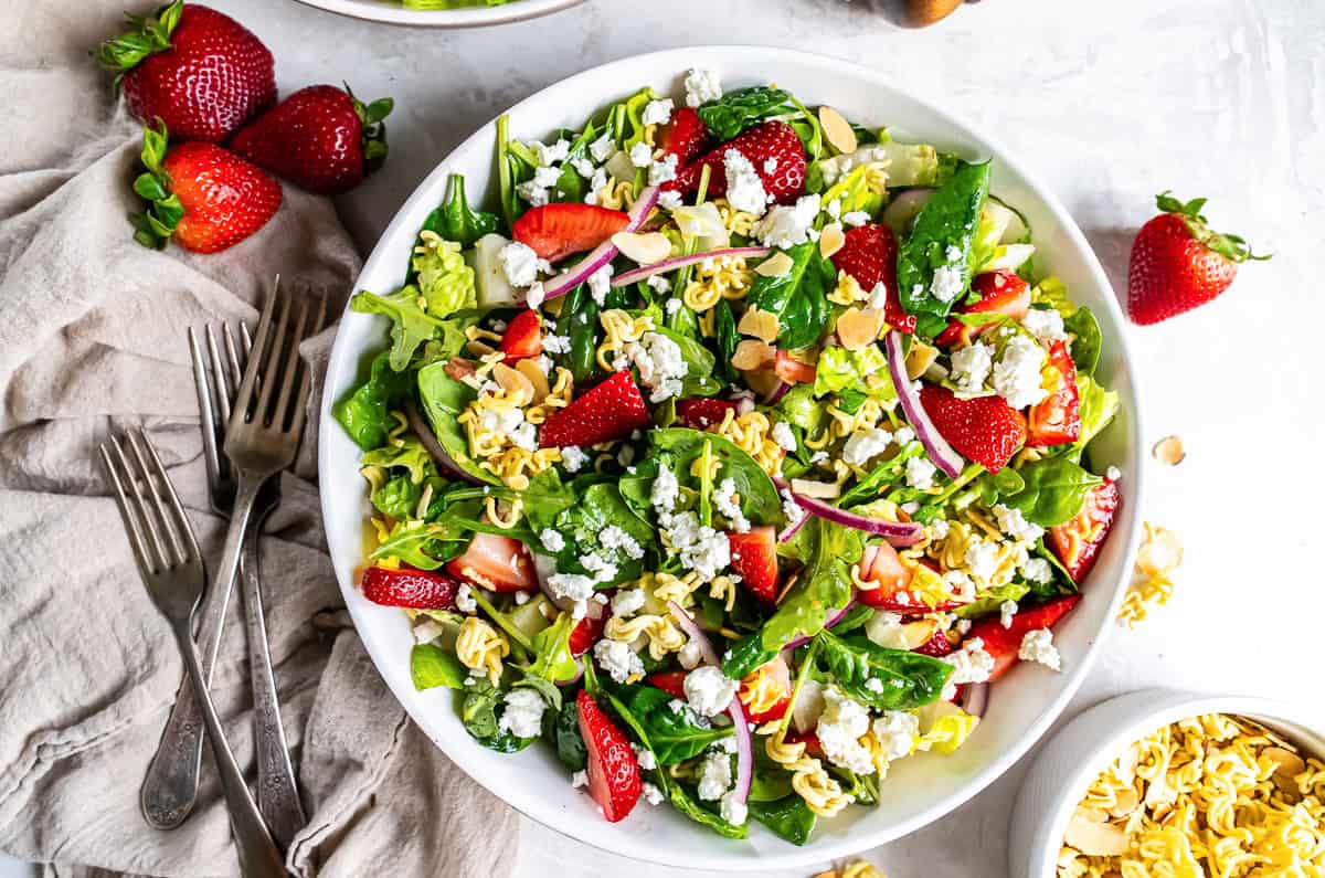 Green salad with strawberries, ramen noodles, almonds, goat cheese and champagne vinaigrette in a large white bowl.